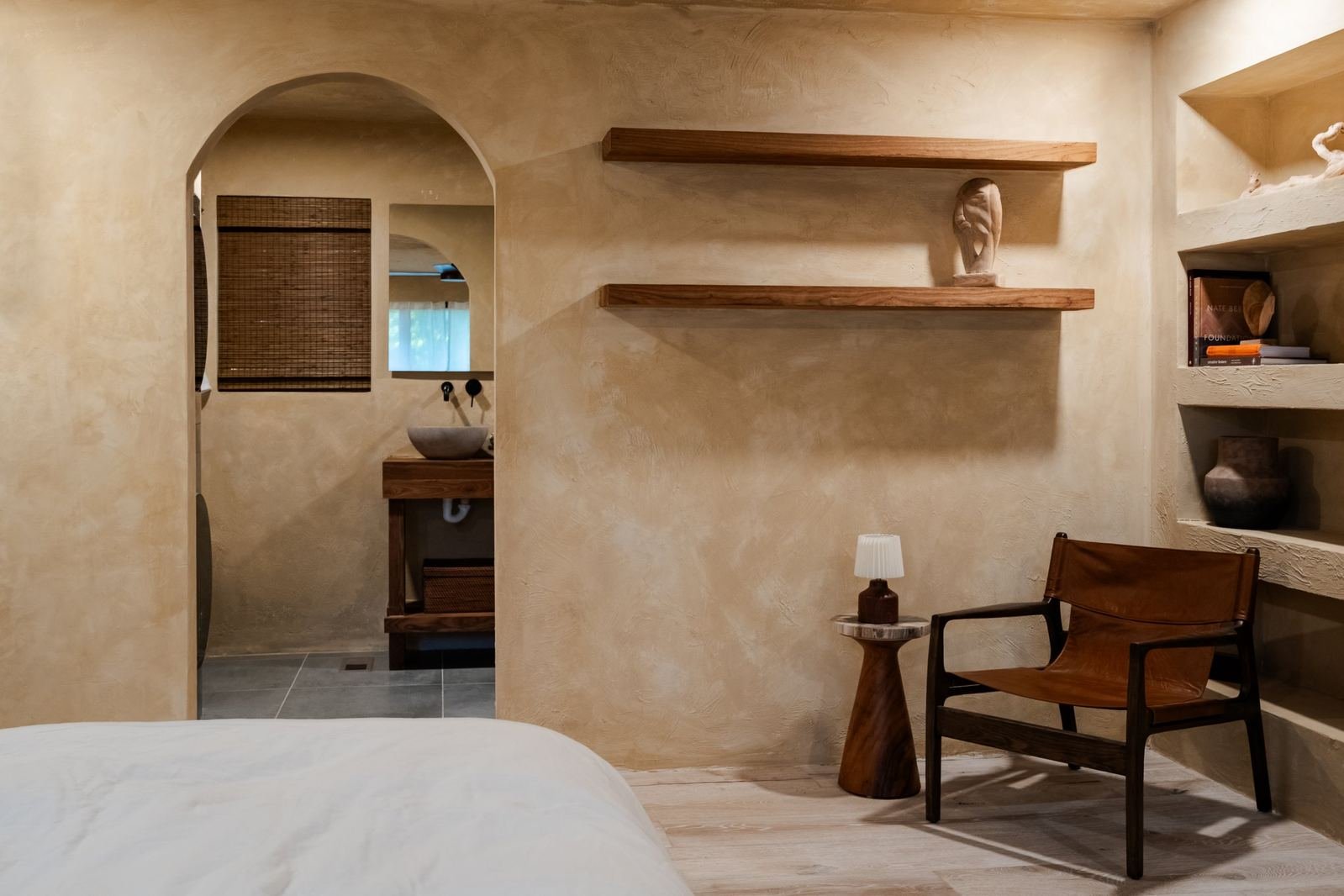 Plaster bedroom with an arched doorway to the bath, floating walnut shelves and a leather sling chair