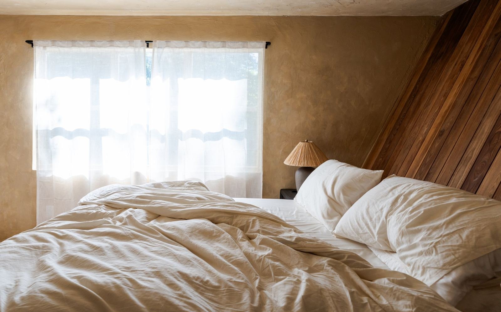 King bed in linen beneath the wood-clad roof pitch, morning light through sheer curtains